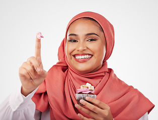 Image showing Portrait, cupcake and icing on finger with a muslim woman in studio on a white background for dessert. Face, smile and food with a happy young islamic person eating a cake, candy or sugar snack