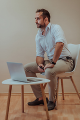 Image showing A confident businessman sitting and using laptop with a determined expression, while a beige background enhances the professional atmosphere, showcasing his productivity and expertise.