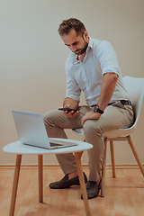 Image showing A confident businessman sitting and using laptop and smartphone with a determined expression, while a beige background enhances the professional atmosphere, showcasing his productivity and expertise.