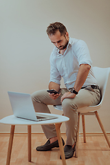 Image showing A confident businessman sitting and using laptop and smartphone with a determined expression, while a beige background enhances the professional atmosphere, showcasing his productivity and expertise.