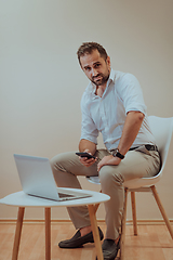 Image showing A confident businessman sitting and using laptop and smartphone with a determined expression, while a beige background enhances the professional atmosphere, showcasing his productivity and expertise.