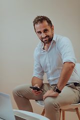 Image showing A confident businessman sitting and using laptop and smartphone with a determined expression, while a beige background enhances the professional atmosphere, showcasing his productivity and expertise.