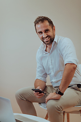 Image showing A confident businessman sitting and using laptop and smartphone with a determined expression, while a beige background enhances the professional atmosphere, showcasing his productivity and expertise.