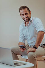 Image showing A confident businessman sitting and using laptop and smartphone with a determined expression, while a beige background enhances the professional atmosphere, showcasing his productivity and expertise.