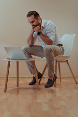 Image showing A confident businessman sitting and using laptop with a determined expression, while a beige background enhances the professional atmosphere, showcasing his productivity and expertise.