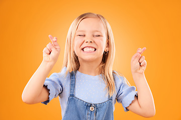 Image showing Child, fingers crossed and wish or hope in studio for luck, prayer or faith. Face of excited young girl kid on a orange background for hand gesture, icon or sign for dream, optimistic or superstition