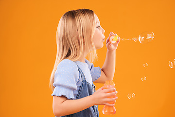 Image showing Girl, child and blowing bubbles in studio for fun, games and childhood development on mockup orange background. Profile of kid playing with soap bubble wand, toys and breathing activity for freedom