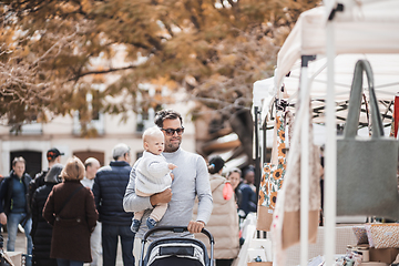 Image showing Father walking carrying his infant baby boy child and pushing stroller in crowd of people wisiting sunday flea market in Malaga, Spain