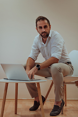 Image showing A confident businessman sitting and using laptop with a determined expression, while a beige background enhances the professional atmosphere, showcasing his productivity and expertise.