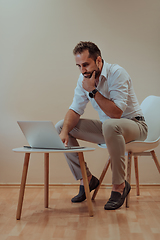 Image showing A confident businessman sitting and using laptop with a determined expression, while a beige background enhances the professional atmosphere, showcasing his productivity and expertise.