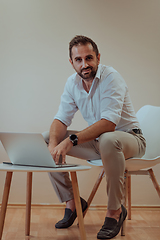 Image showing A confident businessman sitting and using laptop with a determined expression, while a beige background enhances the professional atmosphere, showcasing his productivity and expertise.