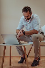 Image showing A confident businessman sitting and using laptop and smartwatch with a determined expression, while a beige background enhances the professional atmosphere, showcasing his productivity and expertise.