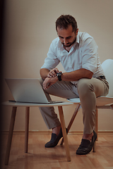 Image showing A confident businessman sitting and using laptop and smartwatch with a determined expression, while a beige background enhances the professional atmosphere, showcasing his productivity and expertise.