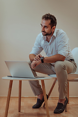 Image showing A confident businessman sitting and using laptop and smartwatch with a determined expression, while a beige background enhances the professional atmosphere, showcasing his productivity and expertise.