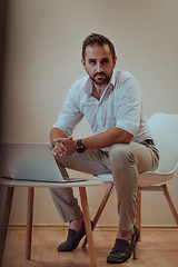 Image showing A confident businessman sitting and using laptop with a determined expression, while a beige background enhances the professional atmosphere, showcasing his productivity and expertise.