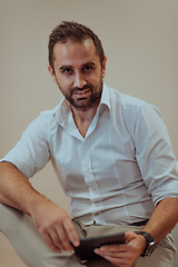 Image showing A confident businessman sitting and using tablet with a determined expression, while a beige background enhances the professional atmosphere, showcasing his productivity and expertise.