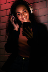 Image showing Woman, phone and headphones for city or night streaming, listening to club music or party in neon light. Happy young person on mobile, social media and audio for techno, rave or network on brick wall