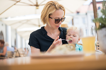 Image showing Young caucasian blonde mother spoon feeding her little infant baby boy child outdoors on restaurant or cafe terrace in summer