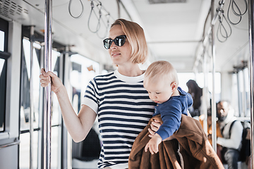 Image showing Mother carries her child while standing and holding on to bar holder on bus. Mom holding infant baby boy in her arms while riding in public transportation. Cute toddler traveling with mother.