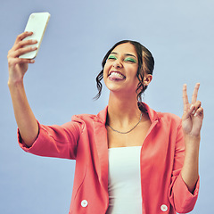 Image showing Phone, fun selfie and woman with a peace sign from social media and tongue out for creative job in studio. Influencer, happy and makeup with career fashion with blue background and emoji hand sign