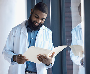 Image showing Doctor, man and reading documents, information or paperwork of patient by window in building with smile. Healthcare, professional or black person check notes, charts on paper with medical experience