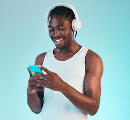 Image showing Headphones, phone and black man in a studio networking and listening to music, playlist or album. Happy, smile and African male model scroll on internet with cellphone and song by blue background.