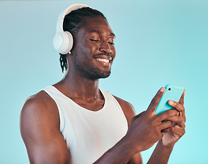 Image showing Headphones, cellphone and black man in a studio networking and listening to music, playlist or album. Happy, smile and African male model scroll on internet with phone and song by blue background.