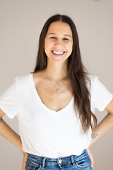 Image showing Portrait of confident beautiful woman with long brown hair, wearing casual clothes, standing in relaxed pose with hands in pockets, smiling with white teeth at camera, studio background.