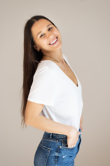 Image showing Portrait of confident beautiful woman with long brown hair, wearing casual clothes, standing in relaxed pose with hands in pockets, smiling with white teeth at camera, studio background.