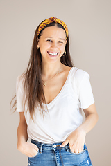 Image showing Portrait of confident beautiful woman with long brown hair, wearing casual clothes, standing in relaxed pose with hands in pockets, smiling with white teeth at camera, studio background.