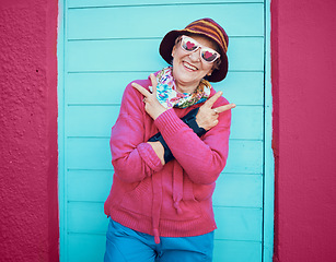 Image showing Portrait, peace and funky with a senior woman outdoor standing against a blue door and red wall background. Glasses, hands and hip hop with a happy mature female doing a hand sign or gesture outside