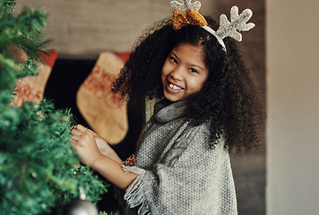 Image showing Christmas, tree and decorating with a girl in antlers putting an ornament on a branch in the living room of her home. Children, portrait and smile with a happy kid placing decorations for celebration