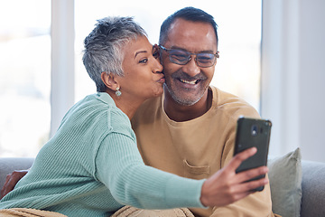 Image showing Phone, selfie and kiss with a senior couple posing for a picture in the living room of their home together. Smartphone, kissing and social media with a mature man and woman taking a photograph