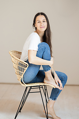 Image showing Portrait of confident beautiful woman with long brown hair, wearing casual clothes, sitting on chair in tight jeans and white t-shirt, studio background