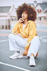 Image showing Trendy, stylish and fashionable female basketball player sitting on a ball at the court against an urban background. Young, edgy and serious woman with an afro posing outside in the city in the day