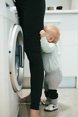 Image showing Little infant baby boy child hiding between mothers legs demanding her attention while she is multitasking, trying to do some household chores in kitchen at home. Mother on maternity leave.