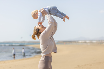 Image showing Mother enjoying summer vacations holding, playing and lifting his infant baby boy son high in the air on sandy beach on Lanzarote island, Spain. Family travel and vacations concept.