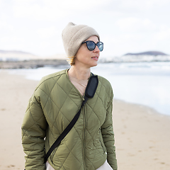 Image showing Portrait of young stylish woman wearing green padded jacket, hoodie, wool cap and sunglasses on long sandy beach in spring.