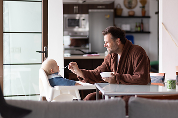 Image showing Father wearing bathrope spoon feeding hir infant baby boy child sitting in high chair at the dining table in kitchen at home in the morning.