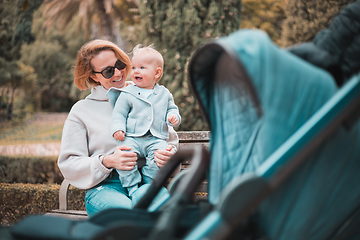 Image showing Mother sitting on bench in urban park, laughing cheerfully, holding her smiling infant baby boy child in her lap having baby stroller parked by their site.