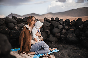 Image showing Mother enjoying winter vacations playing with his infant baby boy son on black sandy volcanic beach of Janubio on Lanzarote island, Spain on windy overcast day. Family travel vacations concept.