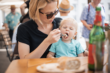 Image showing Young caucasian blonde mother spoon feeding her little infant baby boy child outdoors on restaurant or cafe terrace in summer.