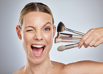 Image showing Woman, face and wink with makeup brushes for cosmetics or facial treatment against a grey studio background. Portrait of female model in beauty holding makeup tools in self care or self love