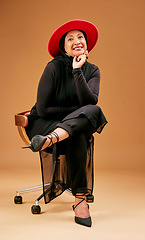 Image showing Fashion, fancy and a mature model woman in studio on a brown background sitting on a chair for trendy clothes. Portrait, hat and clothing with an attractive senior female seated to promote style