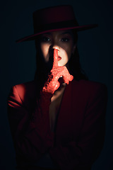 Image showing Fashion, silence and woman in red suit on a dark studio background. Quiet, spotlight and face portrait of beautiful, vintage or 90s female model in makeup cosmetics with shush hand gesture for peace.