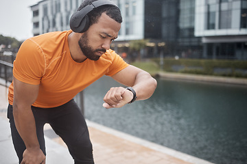 Image showing Fitness, headphones and watch with black man checking running, training and exercise time. Urban, runner and sports athlete listening to music, audio or podcast by water looking at heart rate health