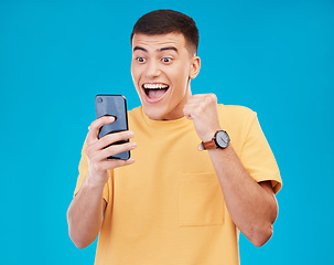 Image showing Cellphone, celebration and young man in a studio with fist pump for winning, success or achievement. Happy, smile and male winner from Canada cheering for prize on a phone isolated by blue background