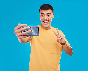 Image showing Man, selfie and peace hand sign with happiness, excited with social media with communication on blue background. V emoji, influencer with memory and smile in picture with photography in a studio