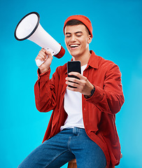 Image showing Phone, megaphone and young man in a studio reading announcement or speech for a rally. Happy, cellphone and male activist or protest with bullhorn for loud communication isolated by blue background