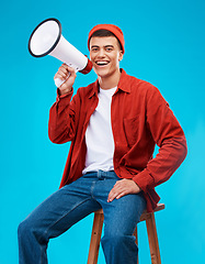 Image showing Portrait, megaphone and young man in a studio for an announcement or speech for a rally. Happy, smile and male activist on stool with bullhorn for loud communication isolated by blue background.